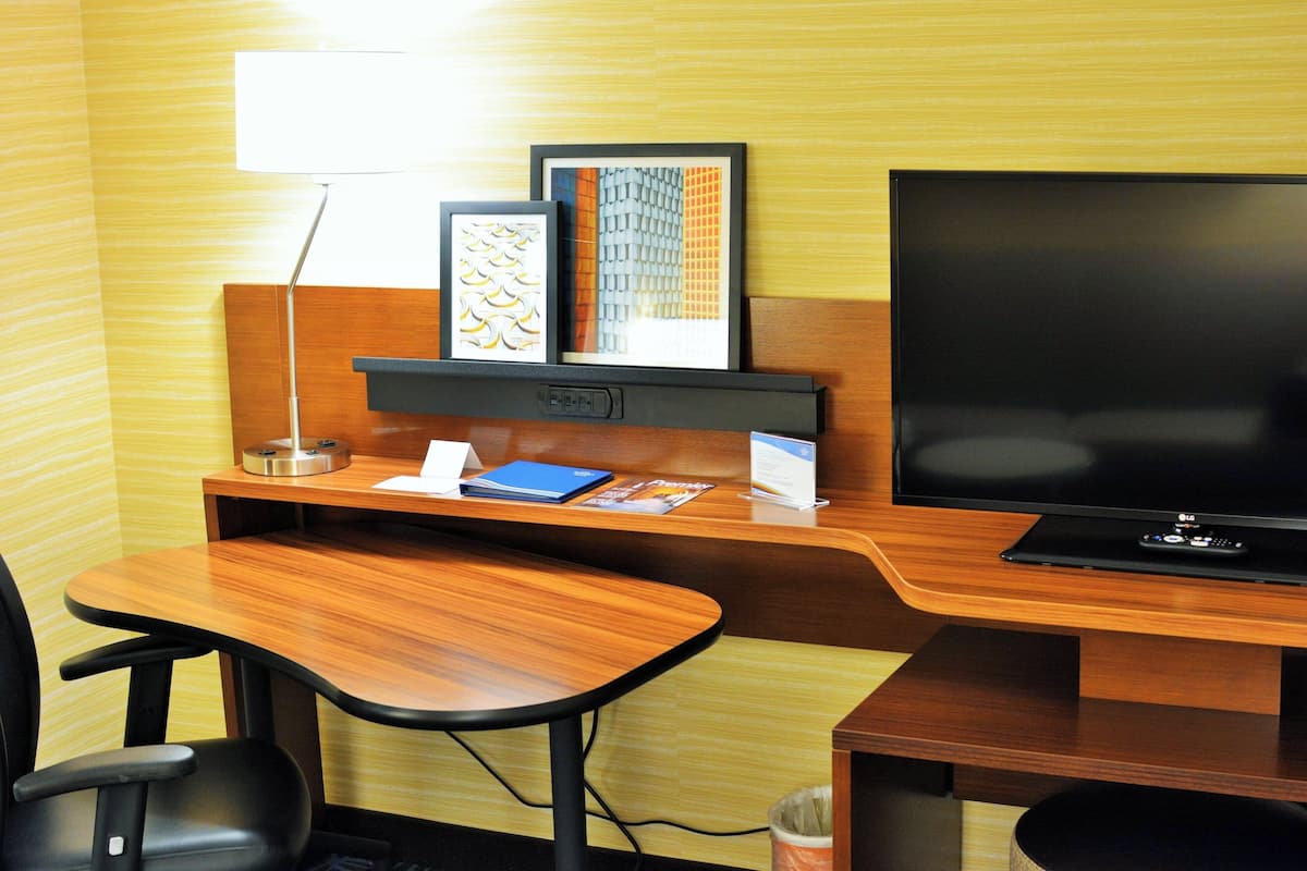 A hotel room with a wooden desk, a television, a lamp, and framed artwork on the wall.