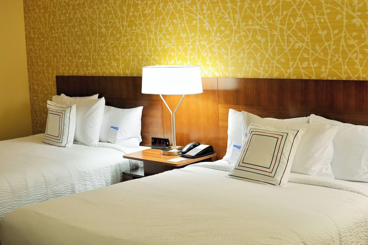 A hotel room with two beds, a nightstand with a lamp, a clock, and a phone.