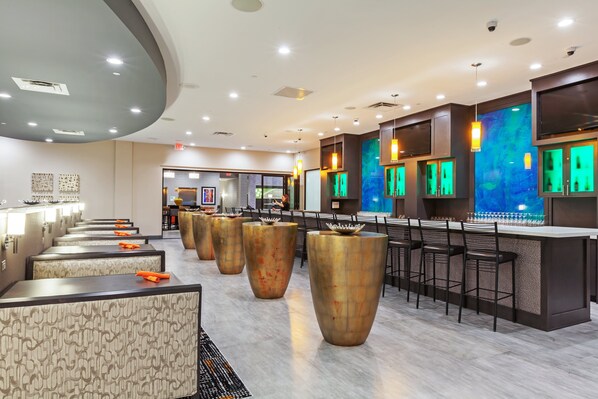 A modern bar area with high stools, a long counter, and a seating area with patterned cushions. - Holiday Inn Houston NE - Bush Airport Area by IHG (Humble)