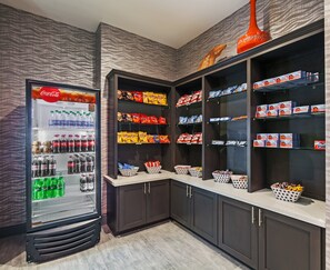A snack and beverage vending area with a Coca-Cola cooler, shelves stocked with chips and snacks, and baskets for additional items. - Holiday Inn Houston NE - Bush Airport Area by IHG (Humble)