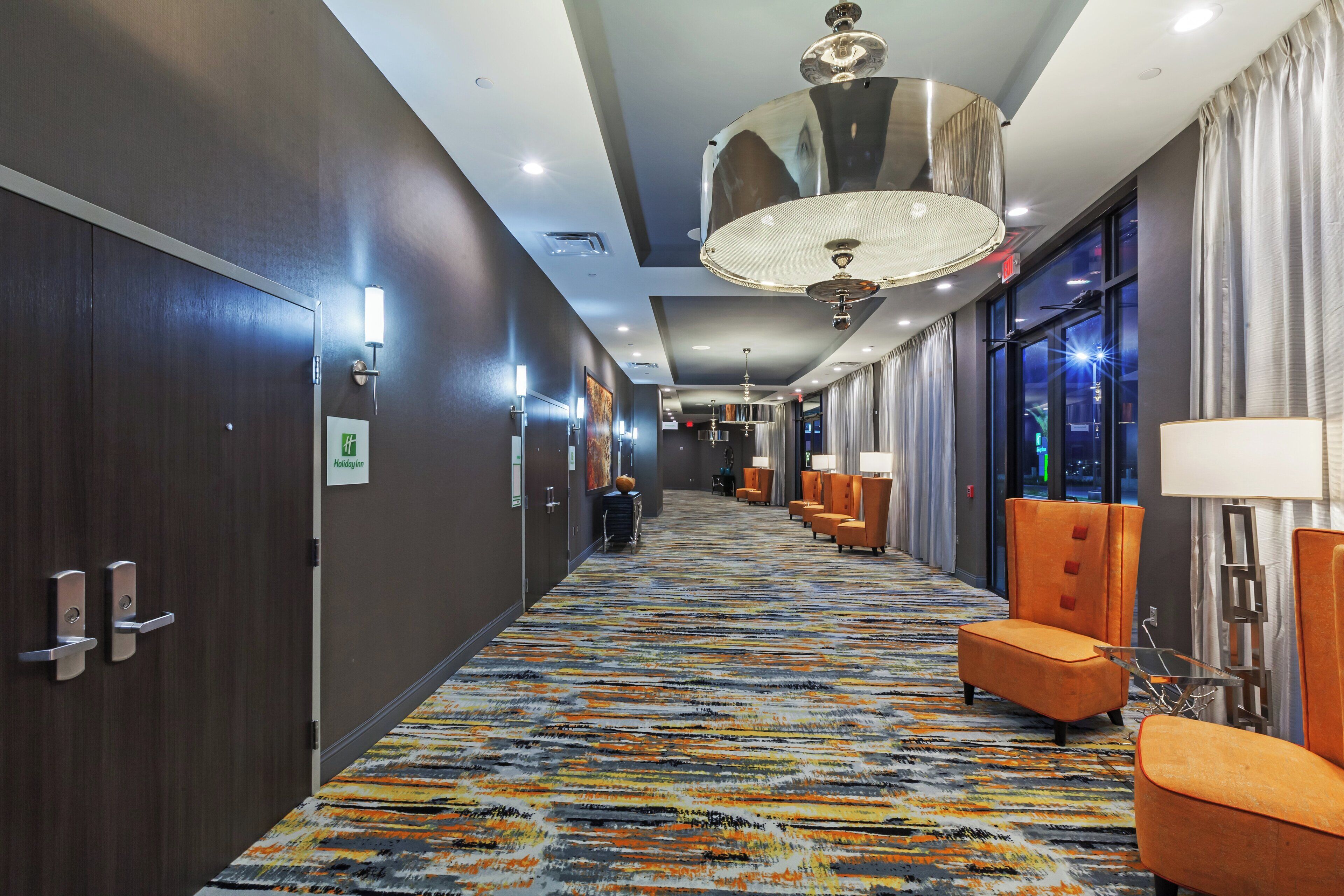 A hotel corridor with a patterned carpet, modern lighting fixtures, and seating areas.