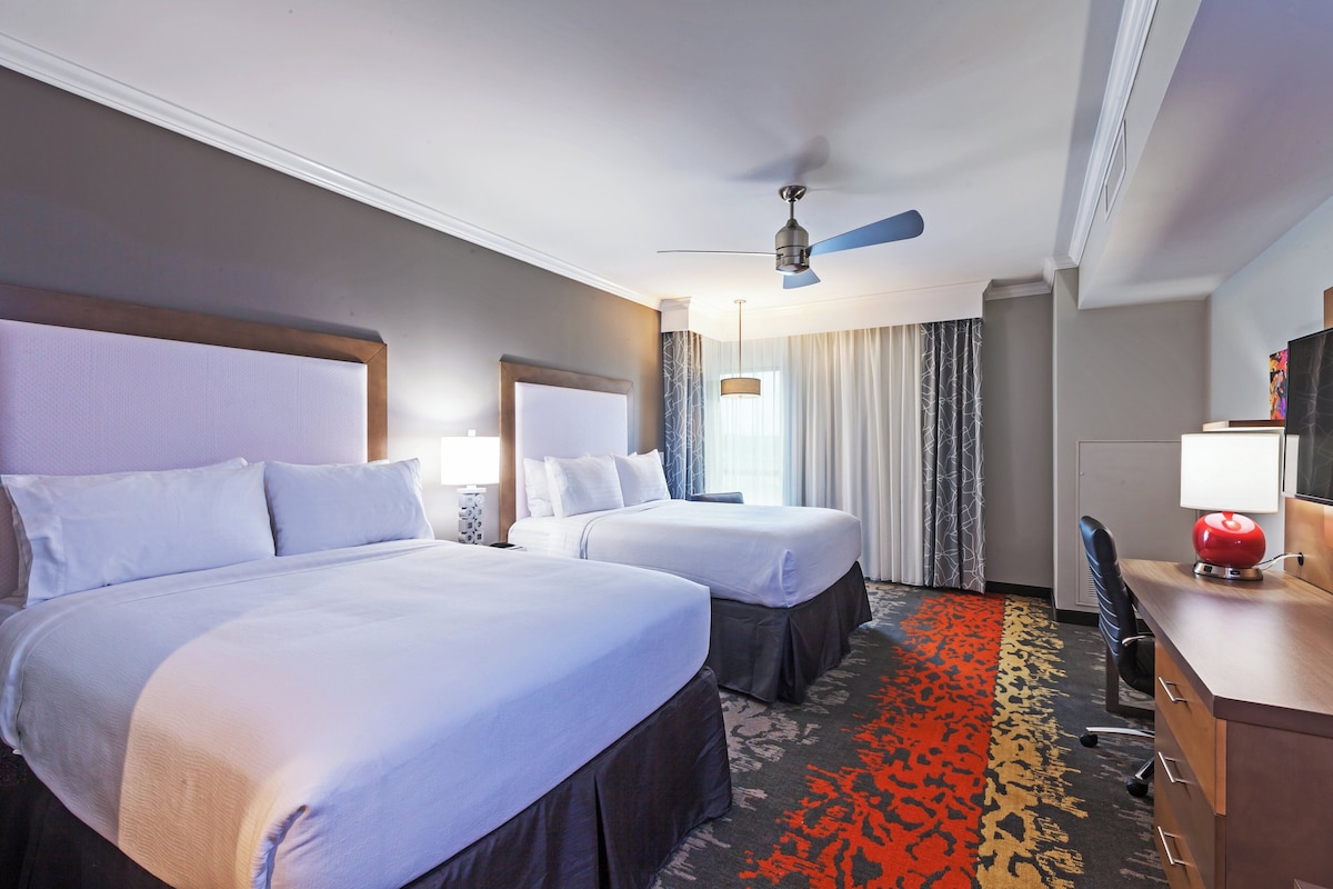 Ein Hotelzimmer mit zwei Betten, einem Schreibtisch, einem Stuhl, einem Deckenventilator und einem großen Fenster mit Vorhängen.