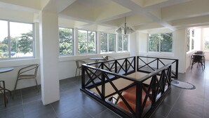 Amplia habitación con ventanas grandes, una zona de estar central con barandilla de madera y un comedor con sillas y mesas.