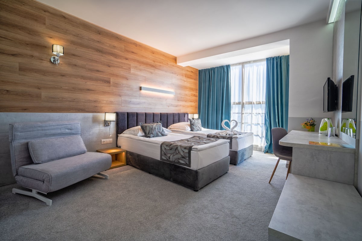 A modern hotel room with a wooden accent wall, a sofa, a bed with a headboard, a desk with a chair, and a TV.