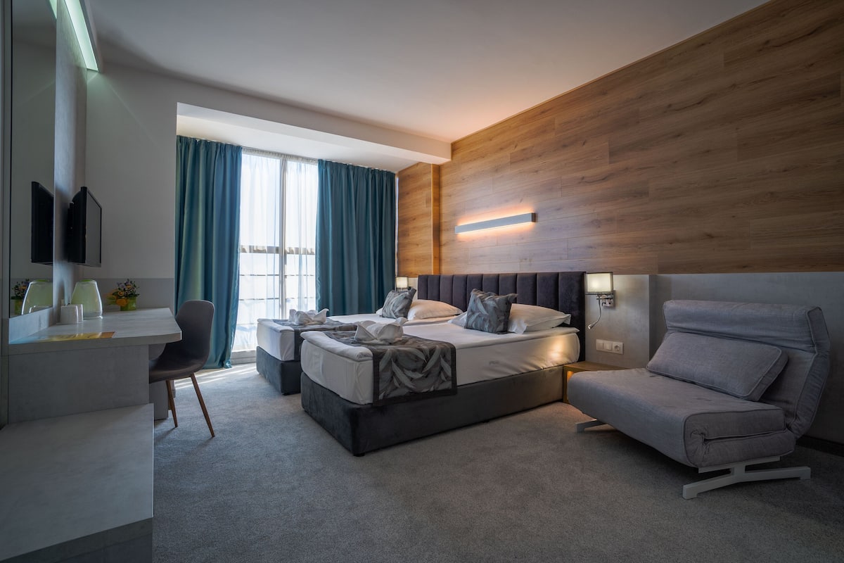 A modern hotel room with a bed, a sofa, a desk, and a large window with curtains.