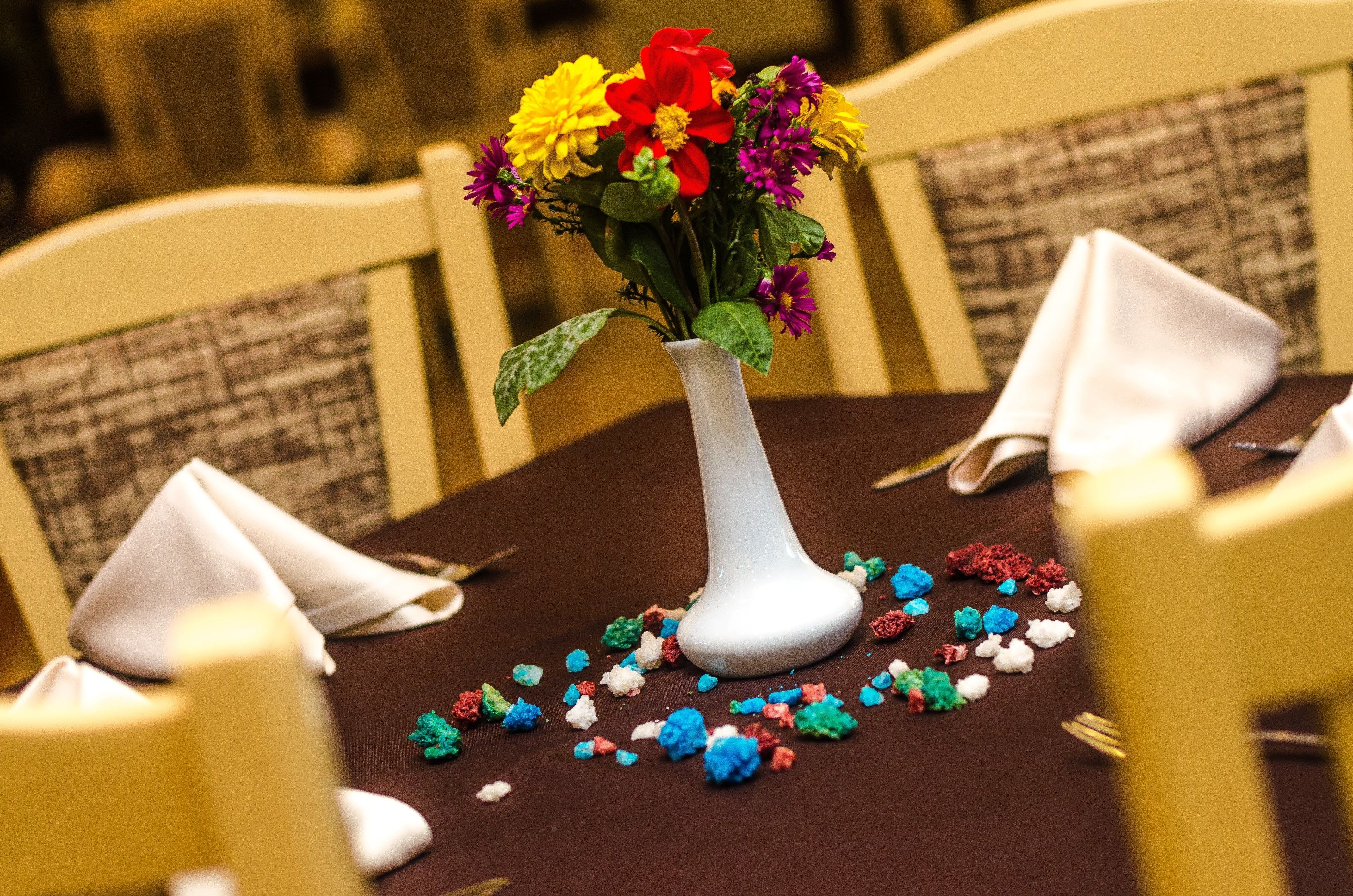 Una mesa de comedor con un jarrón blanco lleno de flores coloridas, servilletas y confeti esparcido.