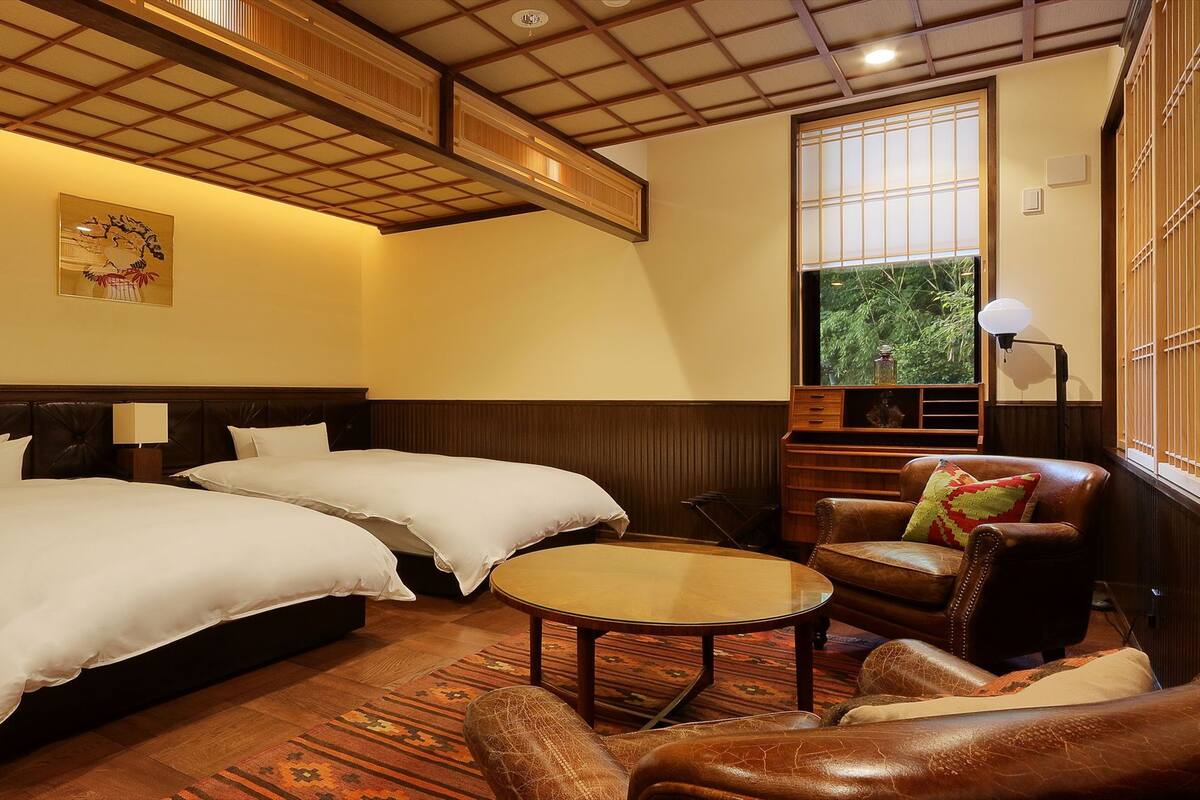 A hotel room with two beds, a small round table, a leather armchair, a wooden cabinet, and a large window with a view of greenery.