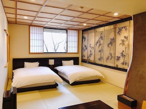 A room with two beds, a large window with a view of trees, and a traditional bamboo screen on the wall.