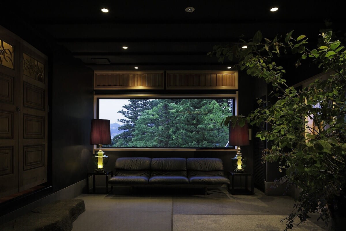 A dark room with a large window, a sofa, two lamps, and a plant.