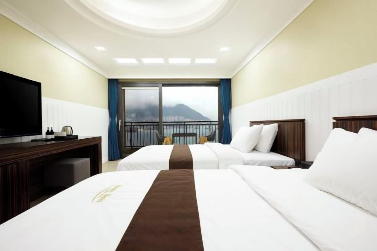 A hotel room with two beds, a TV, a desk, and a balcony with a view of a mountain.