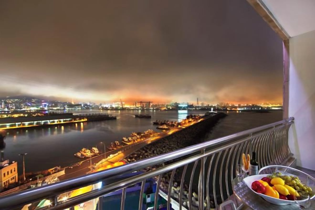 A balcony with a view of a river at night, a cityscape in the background, and a table with fruit and a bottle.