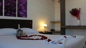 A hotel room with a neatly made bed, a bedside lamp, a bottle of champagne, and a bouquet of flowers on a nightstand. - Nogal Hotel (La Ceja)
