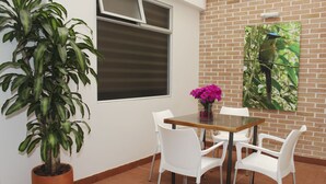 A dining area with a table, four white chairs, a potted plant, a window, and a picture on the wall. - Nogal Hotel (La Ceja)