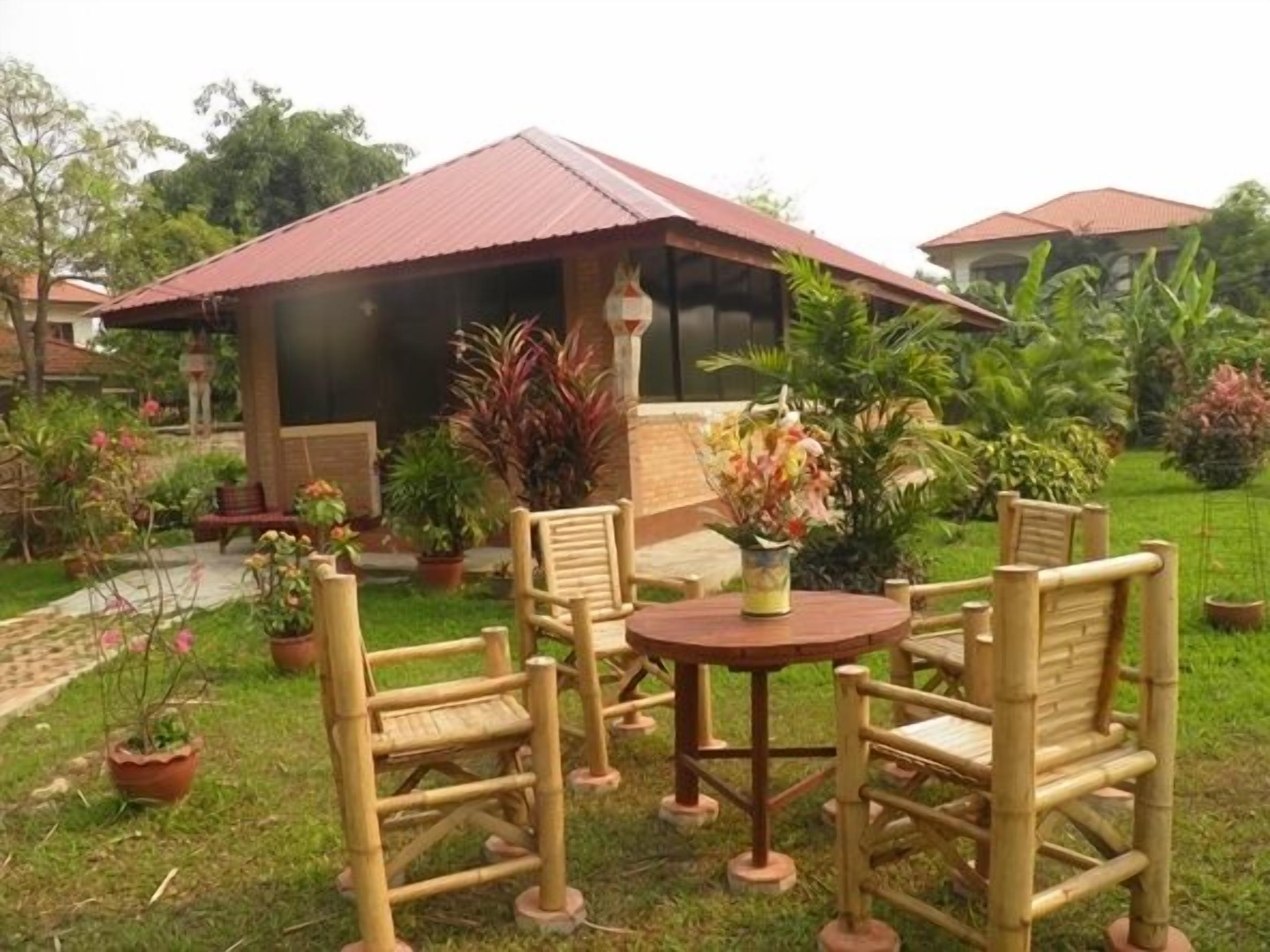 An outdoor seating area with bamboo furniture and a small table, surrounded by potted plants and a building with a red roof.