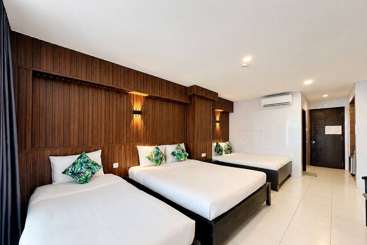 Une chambre d’hôtel avec trois lits, des panneaux de bois au mur, des lampes fixées au mur, un appareil de climatisation et une porte.