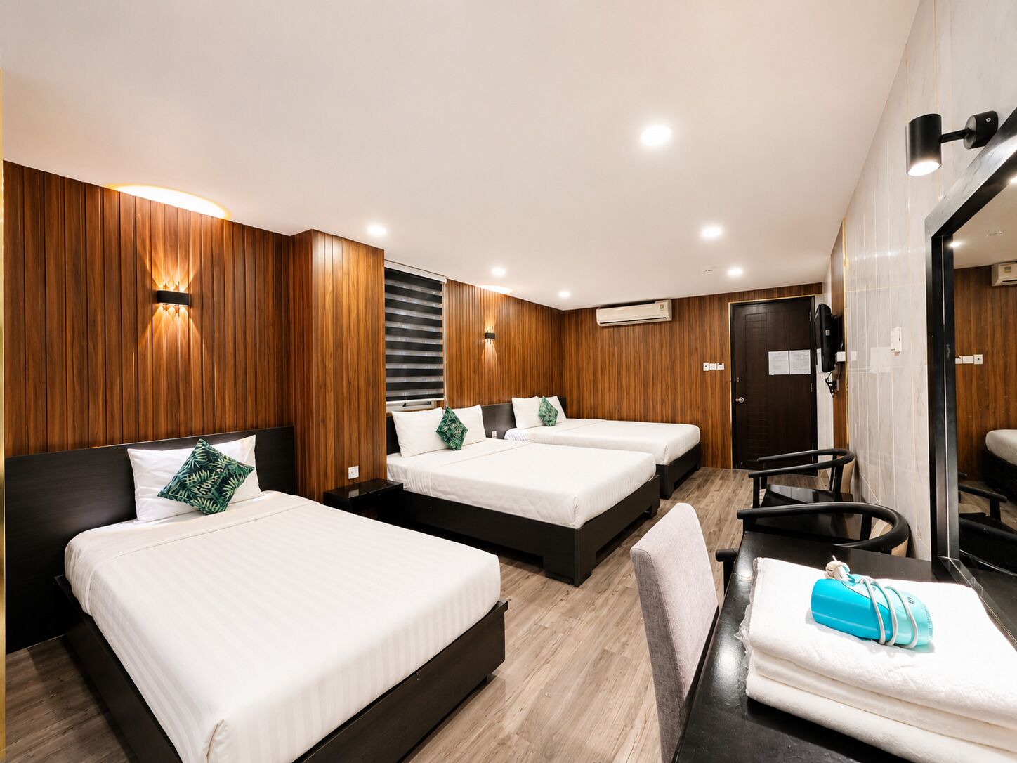 Une chambre d’hôtel avec trois lits, un lambris en bois, un bureau avec une chaise et une salle de bain avec un miroir.
