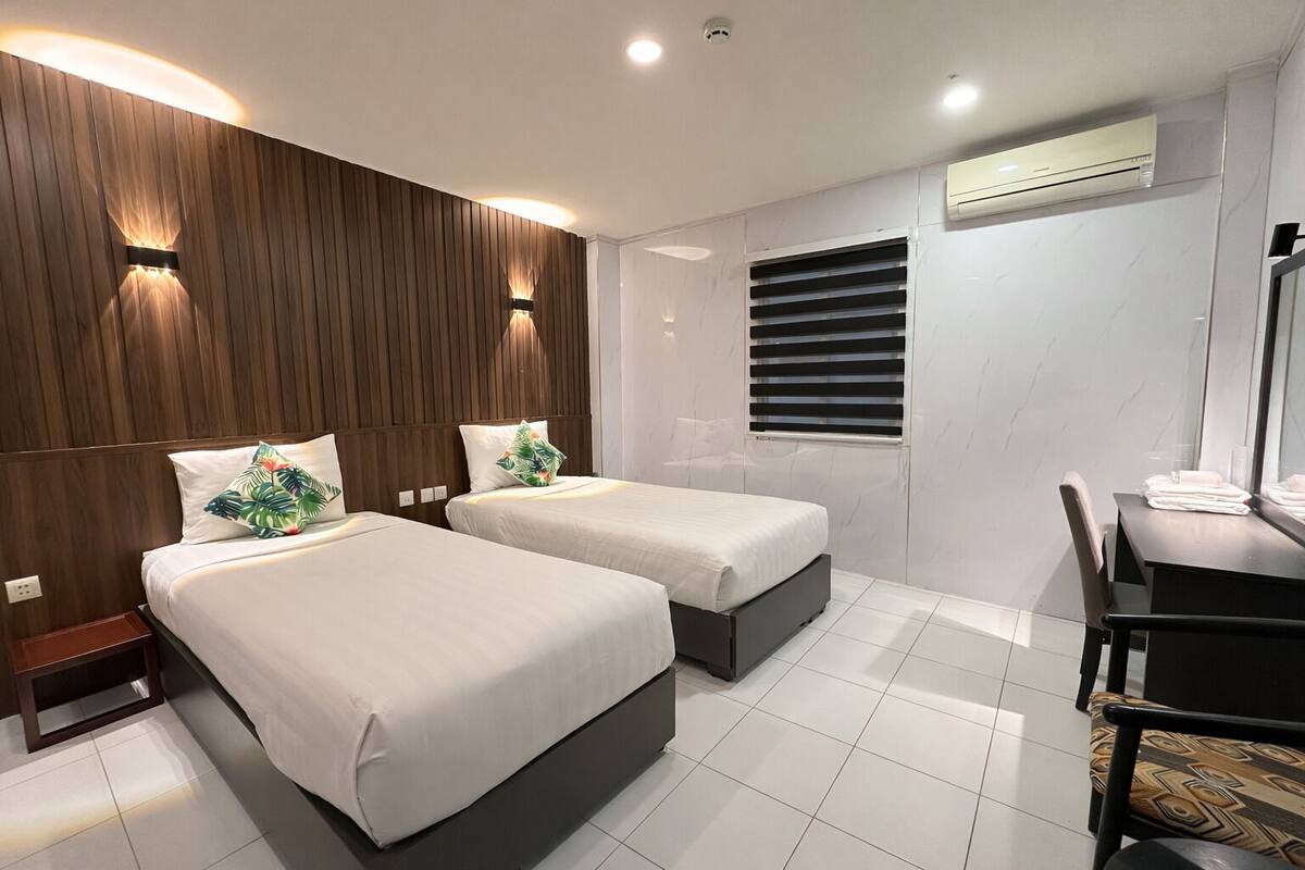 Une chambre d’hôtel avec deux lits, un bureau, une chaise, une fenêtre avec des stores et un appareil de climatisation.