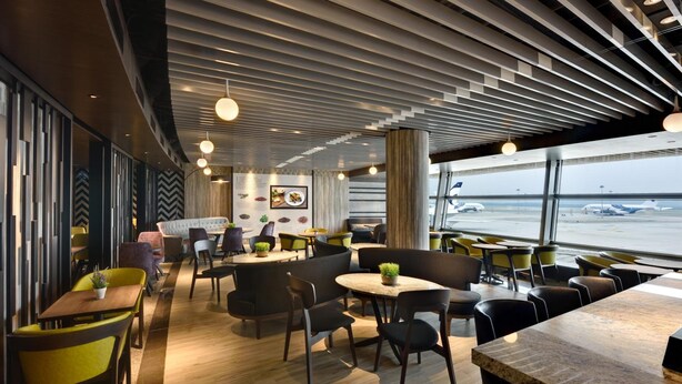 Restaurante moderno com piso de madeira, cadeiras pretas e amarelas, e vista para o aeroporto.
