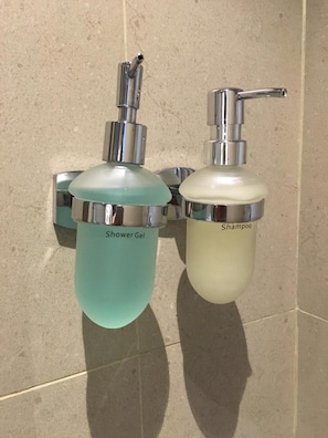 Twee wandmontage doseerders, één met de tekst 'Shower Gel' en de ander 'Shampoo', met een betegelde achtergrond.