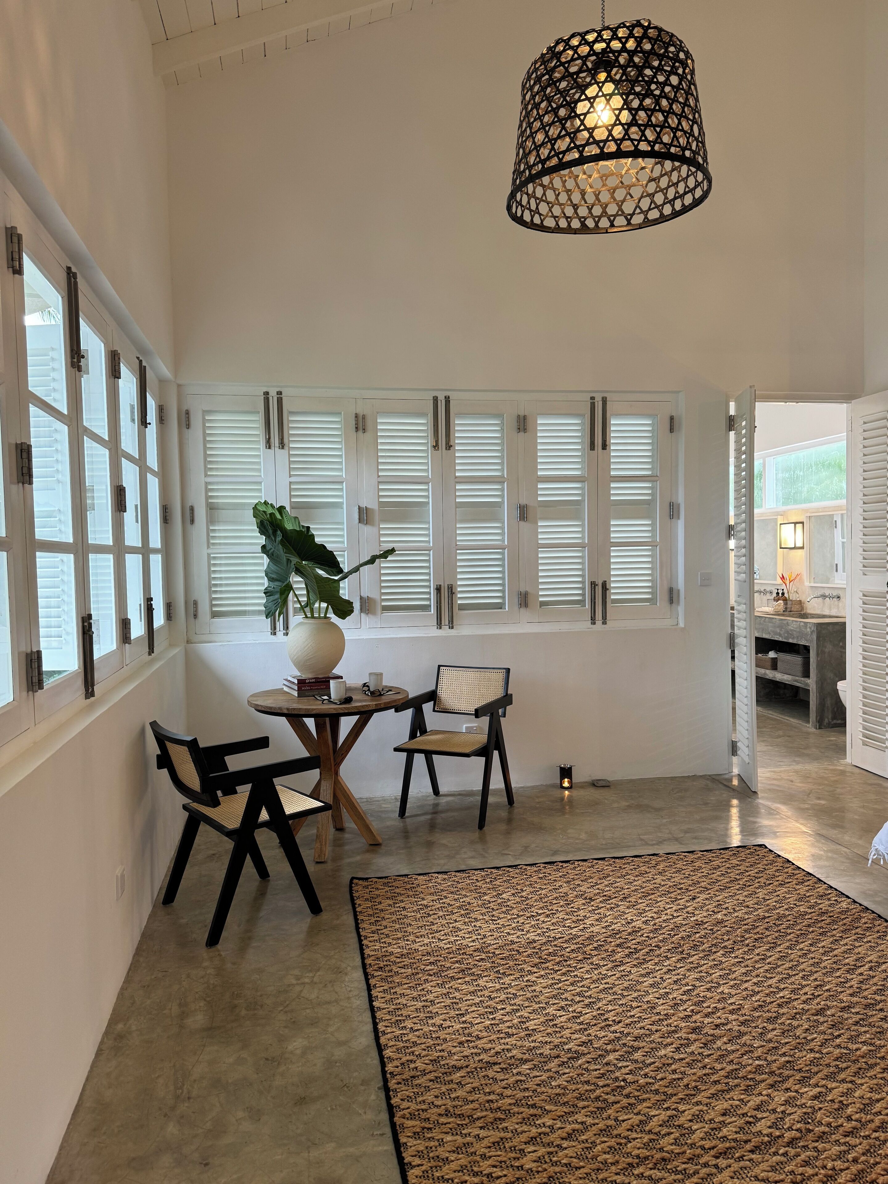 A spacious room with a large window, a dining table with chairs, a potted plant, and a hanging light fixture.