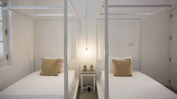 A room with two beds, each with white bedding and beige pillows, a small table with water bottles, and a hanging light fixture.