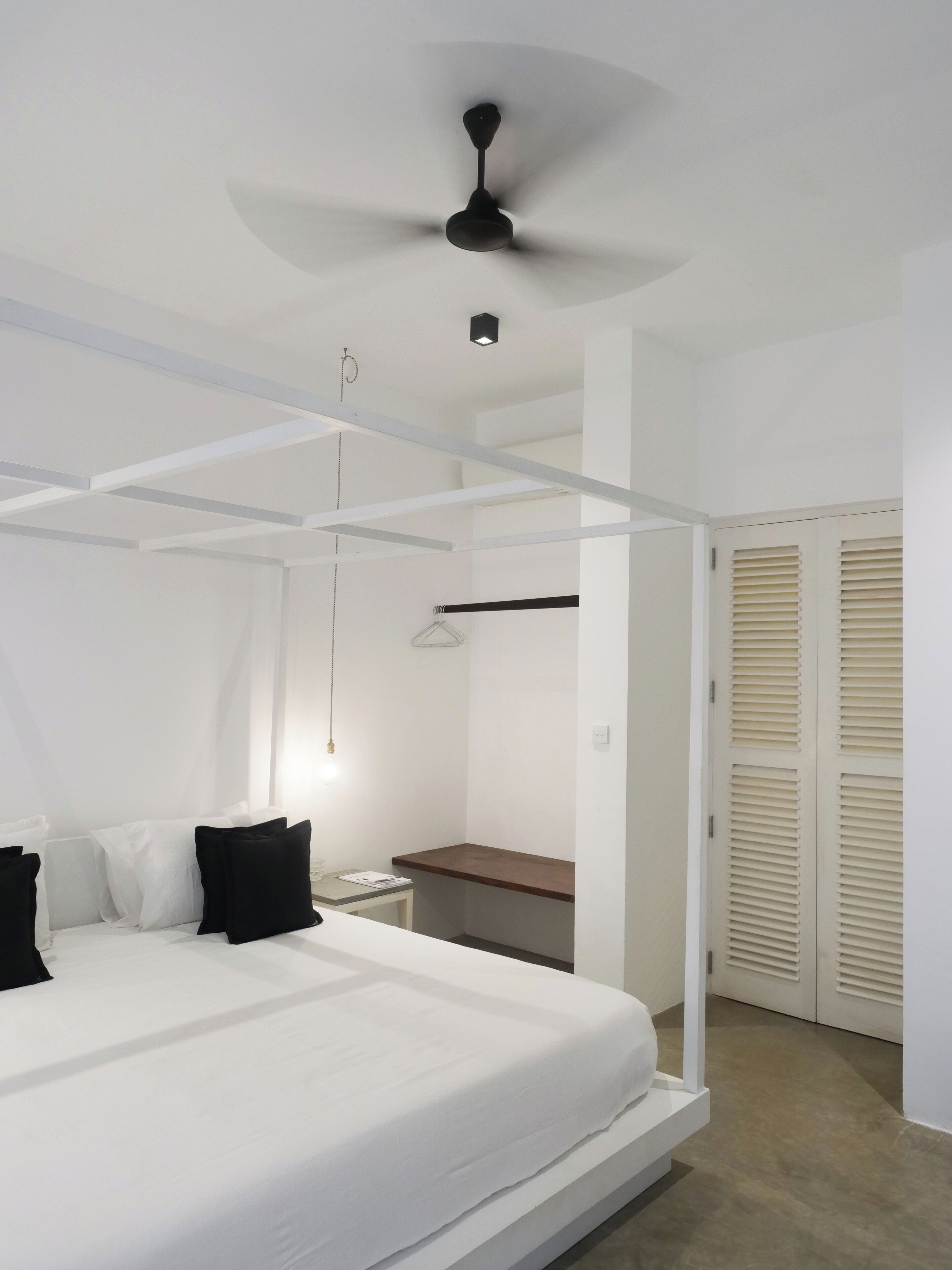 A bedroom with a four-poster bed, a ceiling fan, a bedside table, and a wardrobe with louvered doors.