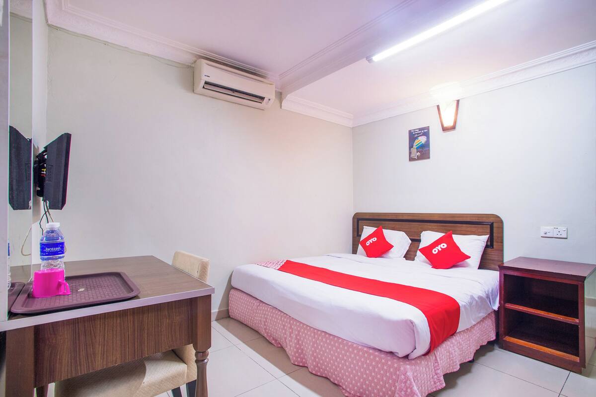 A hotel room with a bed, a desk, a chair, a nightstand, and an air conditioning unit.