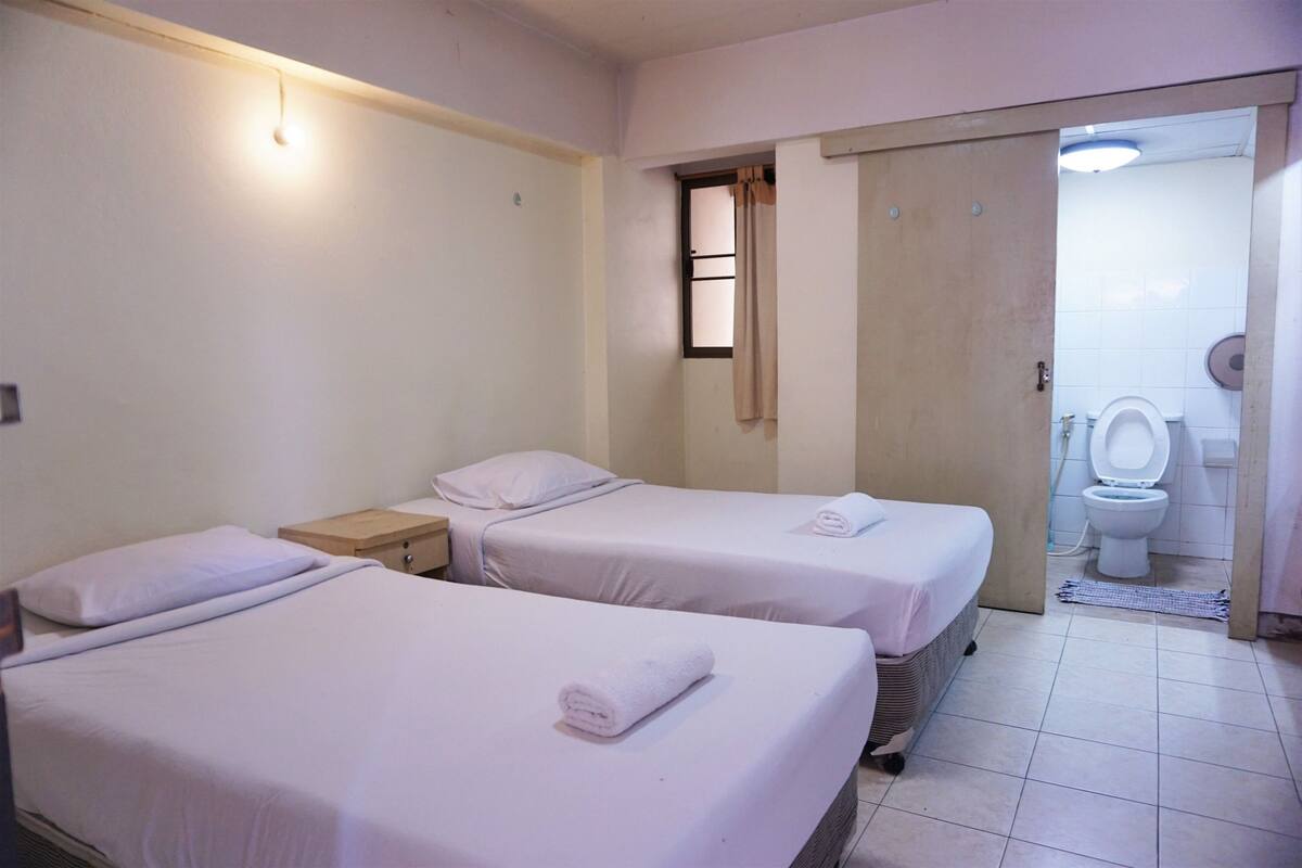 A hotel room with two beds, a small table, a window with curtains, and a bathroom visible through an open door.