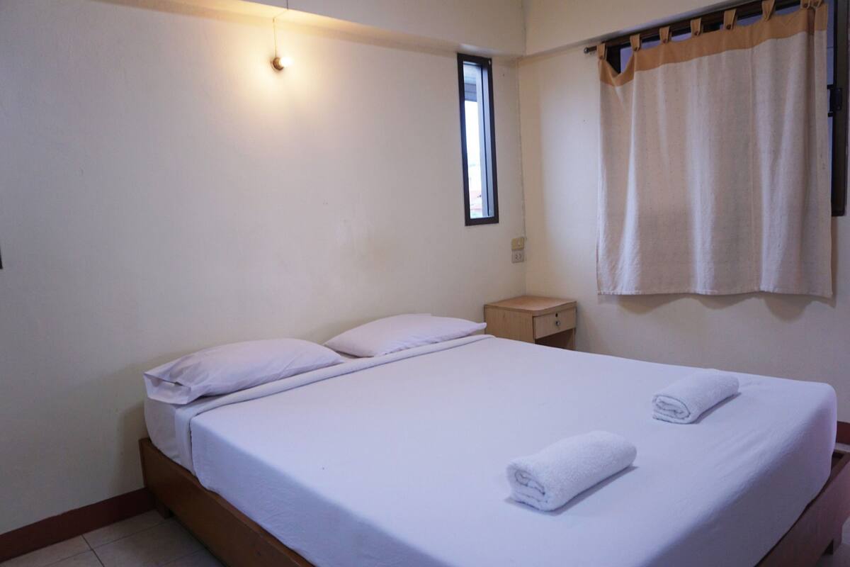 A single bed with white linens and two rolled towels, a small bedside table, a window with a curtain, and a wall-mounted light fixture.