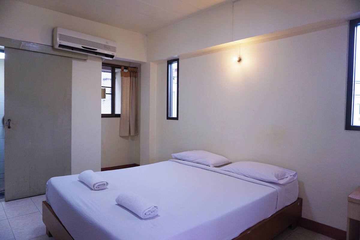 A single bed with white linens, a bedside table, a wall-mounted air conditioner, and a window with curtains.