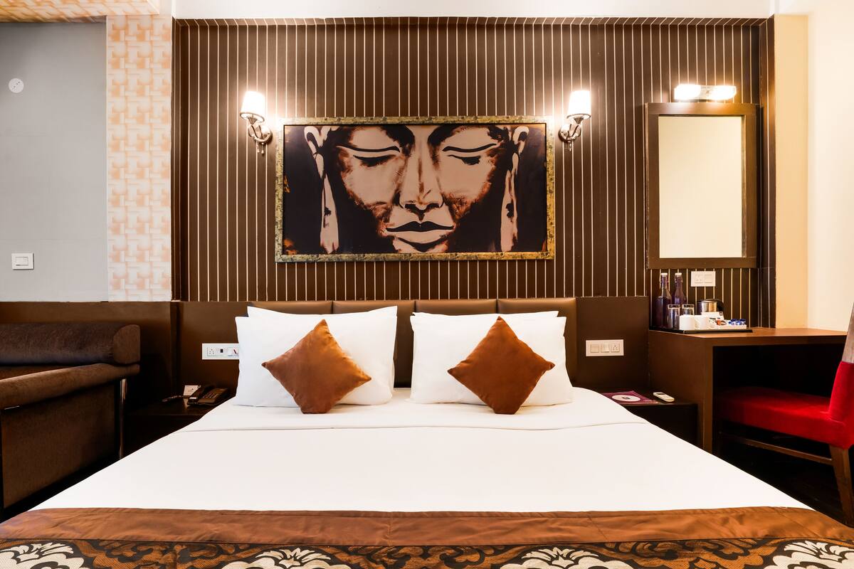 Une chambre d’hôtel avec un grand lit, deux oreillers marron, une tête de lit, une table de chevet avec une lampe, une chaise rouge, un miroir et une œuvre d’art accrochée au mur.