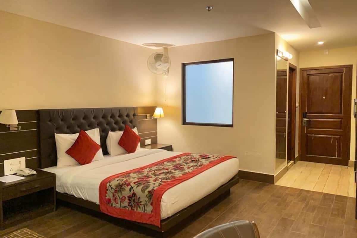 Ein Hotelzimmer mit einem großen Bett, einem Nachttisch mit Lampe, einem an der Wand montierten Fernseher und einer Tür zu einem weiteren Raum.