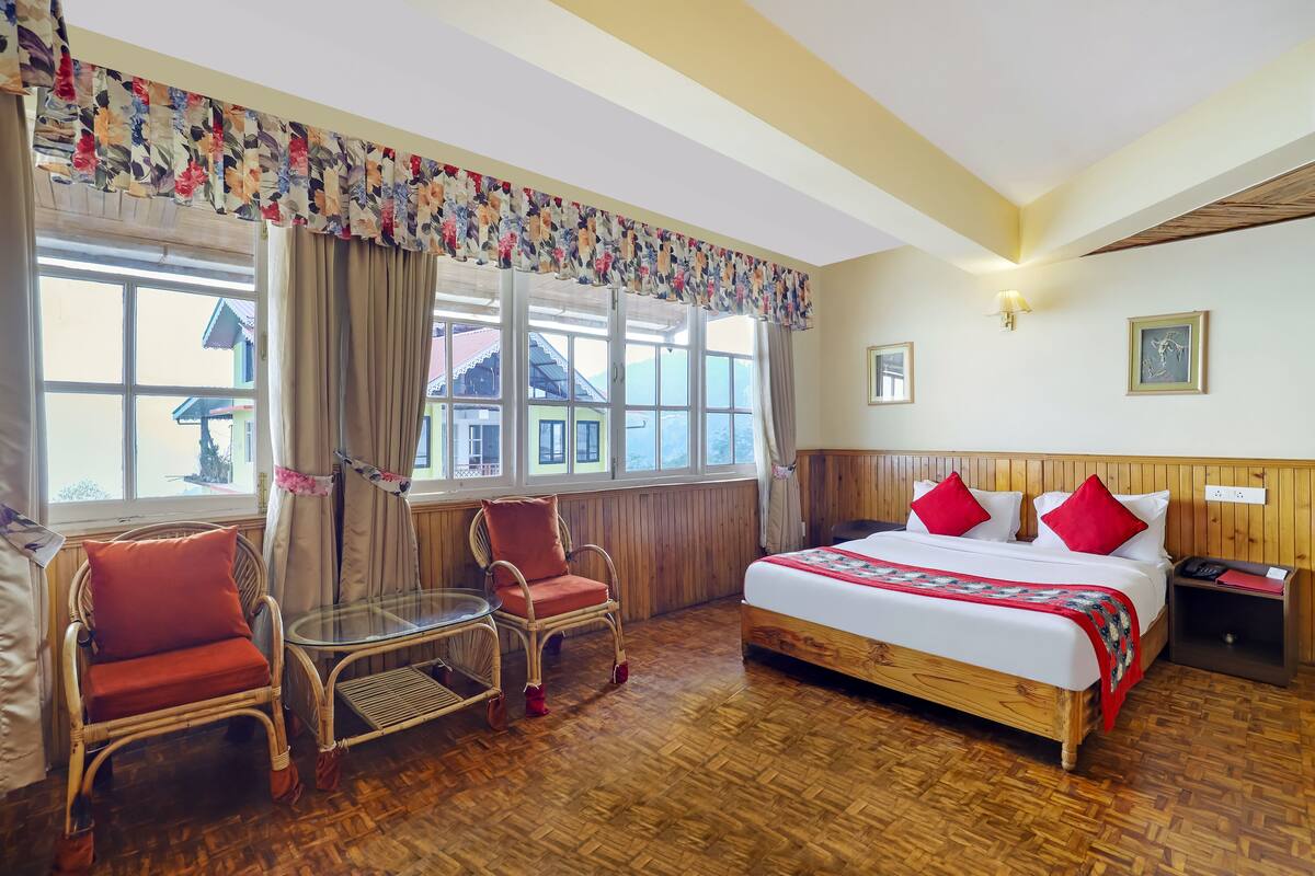 Une chambre d’hôtel avec un plancher en bois, un lit avec des oreillers rouges, une chaise en osier, une table en verre et une fenêtre avec des rideaux.