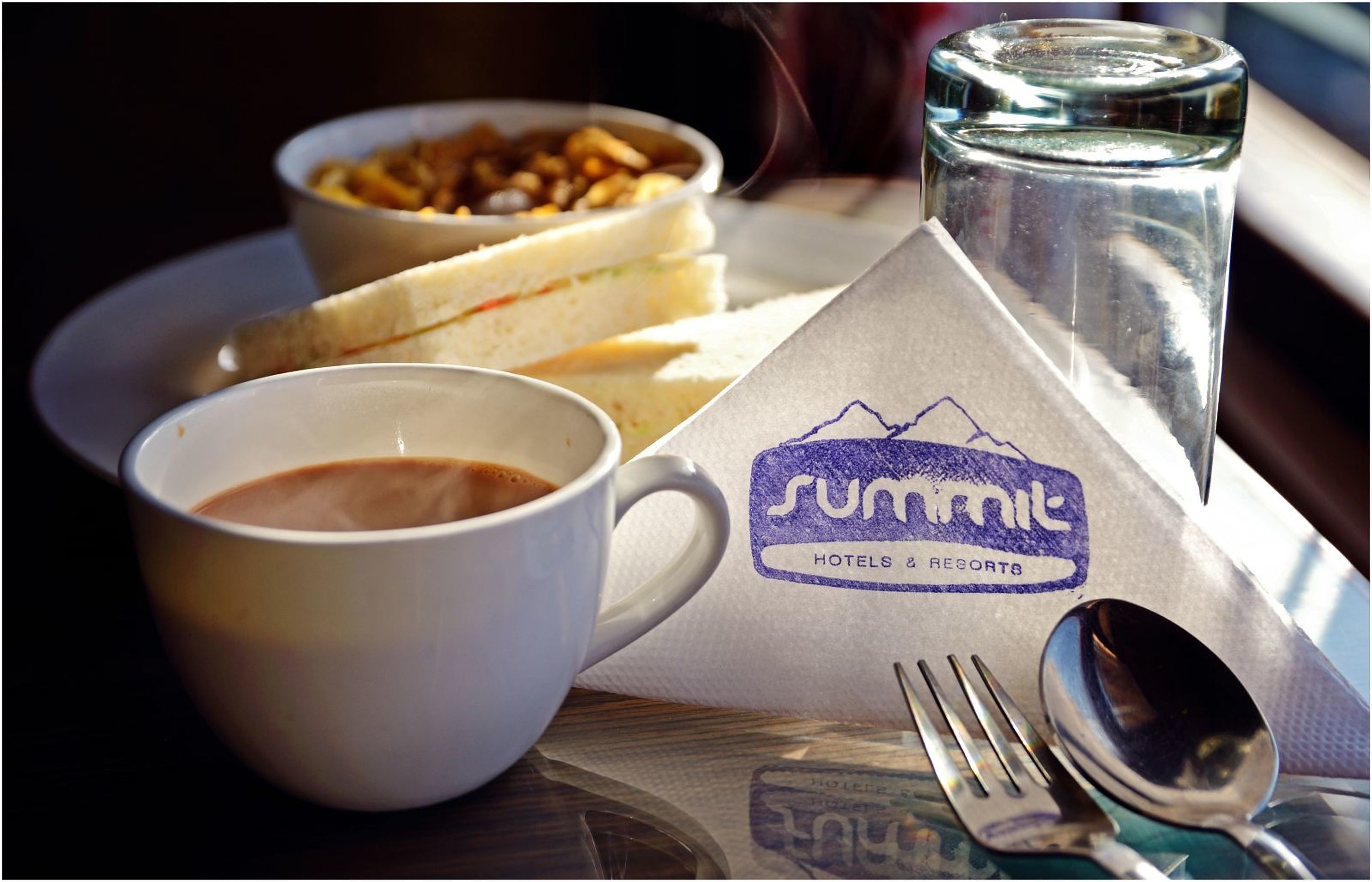 Ein Tablett mit einem Sandwich, einer Nussmischung, einer Tasse Kaffee und einem Glas Wasser, sowie einem Servietten und Besteck.
