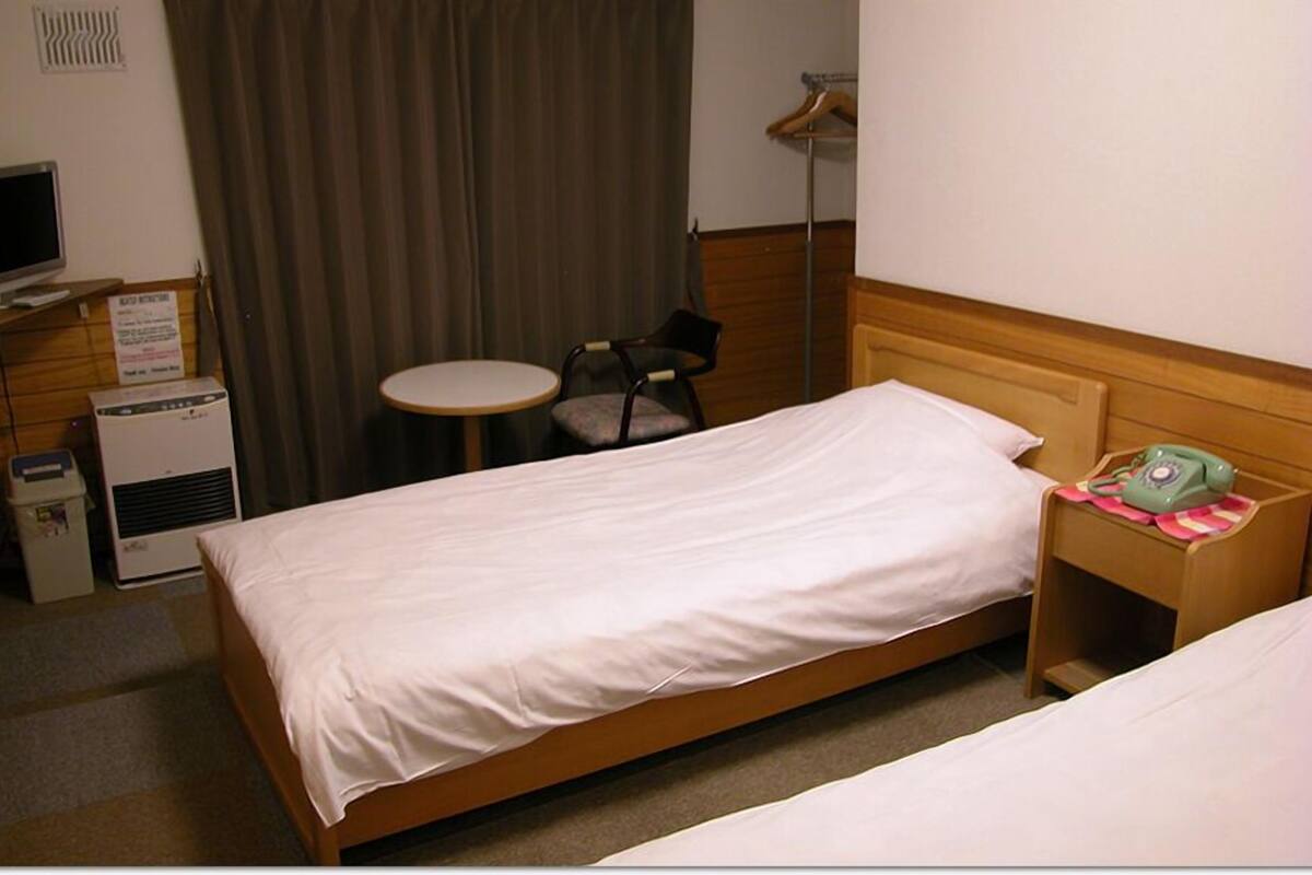 A hotel room with a single bed, a bedside table with a telephone, a chair, a small round table, and a television.