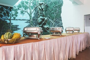 A buffet table with food warmers and a fruit basket, set against a mural of a tropical landscape. - Neptune Bay Hotel (Unawatuna)