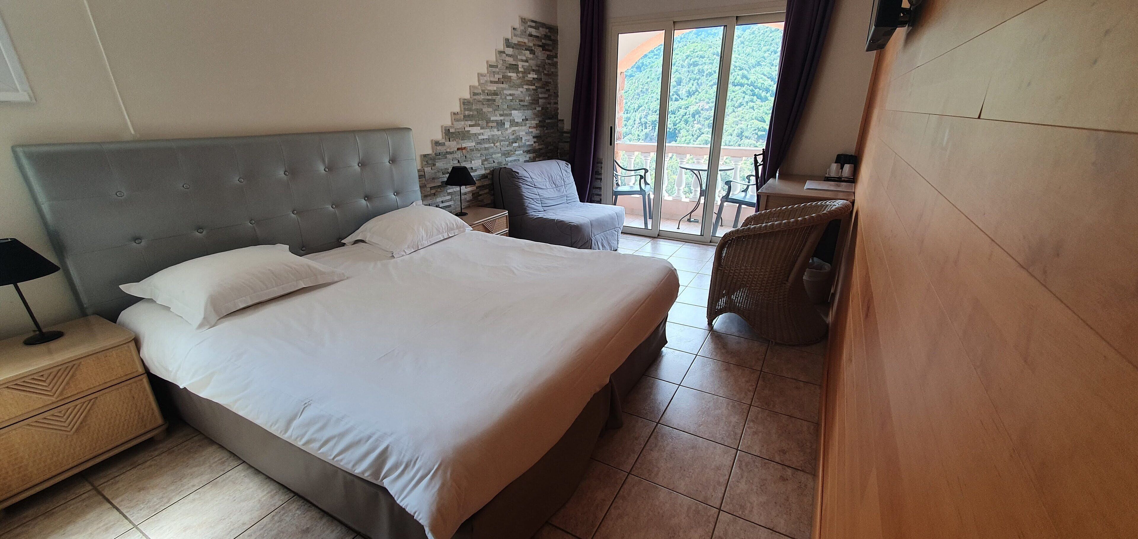 A hotel room with a large bed, a nightstand, a chair, and a view of a balcony with outdoor furniture.