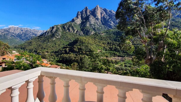 Un balcón con vistas a un paisaje montañoso, una carretera con coches y edificios con tejados rojos.