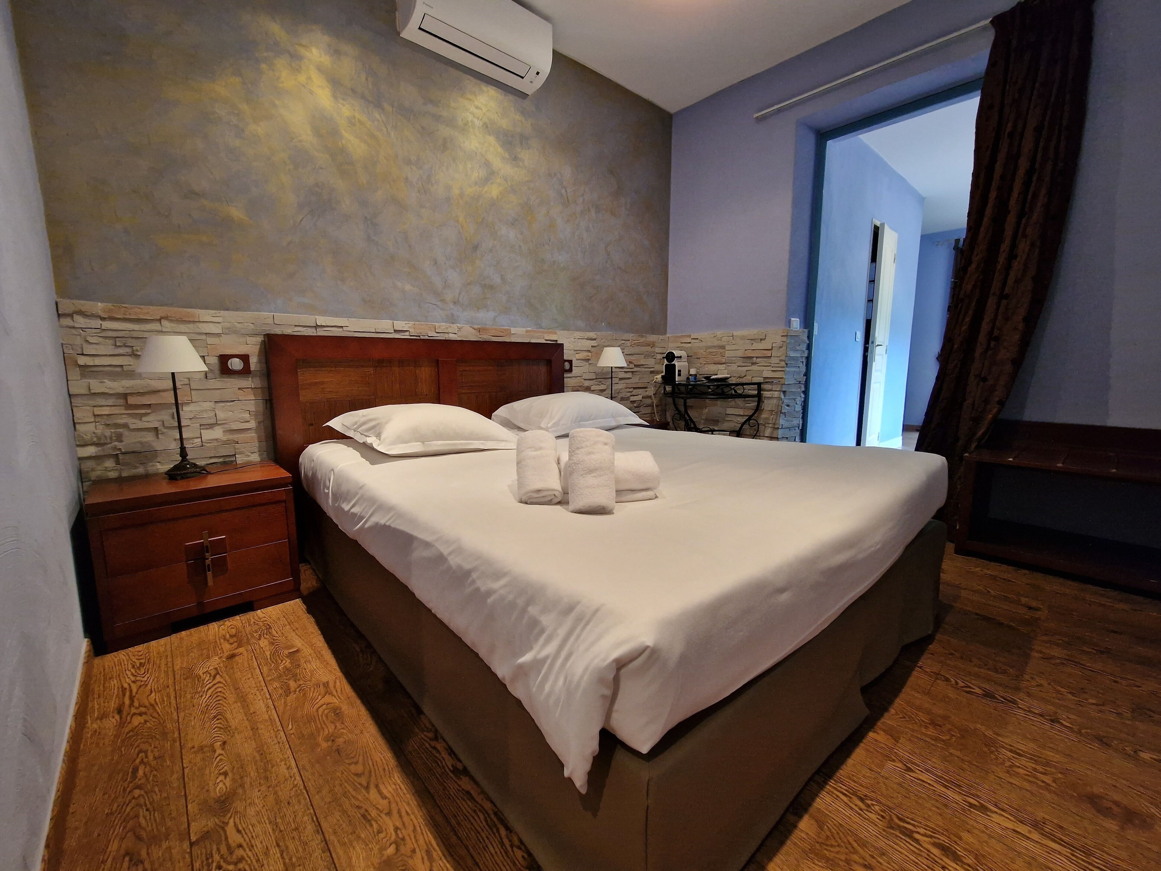 Un lit bien fait, avec du linge de lit et des serviettes blanches, une table de chevet en bois avec une lampe, un mur à la texture de pierre et une porte donnant sur l’extérieur.