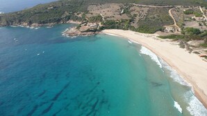 Vista aérea de una zona costera con playa arenosa, aguas turquesas cristalinas y un escarpado telón de fondo montañoso.