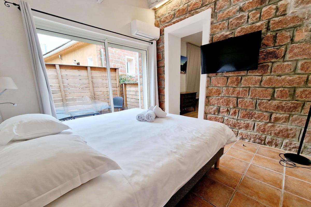 Una cama bien hecha con ropa blanca, un televisor de pantalla plana montado en una pared de ladrillo y una ventana con cortinas.