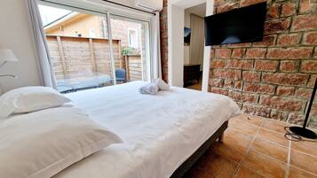 A neatly made bed with white linens, a flat-screen TV mounted on a brick wall, and a window with curtains.