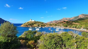 Un pueblo costero con una estructura destacada en la cima de una colina, un puerto deportivo con barcos y abundante vegetación.