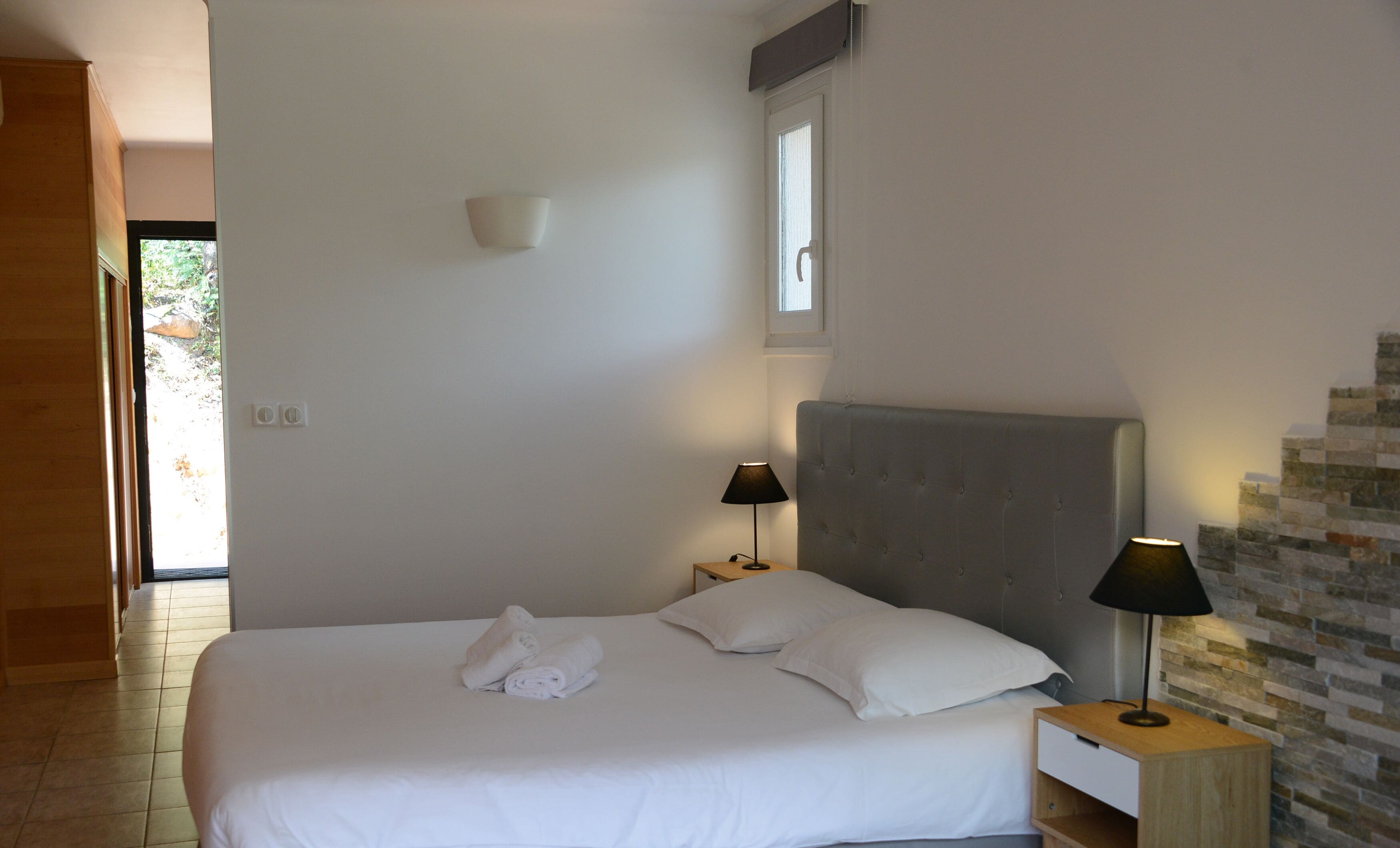 A neatly made bed with white linens and towels, a grey upholstered headboard, two bedside lamps, and a small table with a lamp.