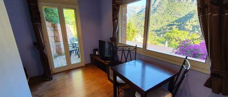 Ein Zimmer mit Holzboden, einem Tisch mit Stühlen, einem Fernseher auf einem Schrank und einem großen Fenster mit Blick auf die Berge.