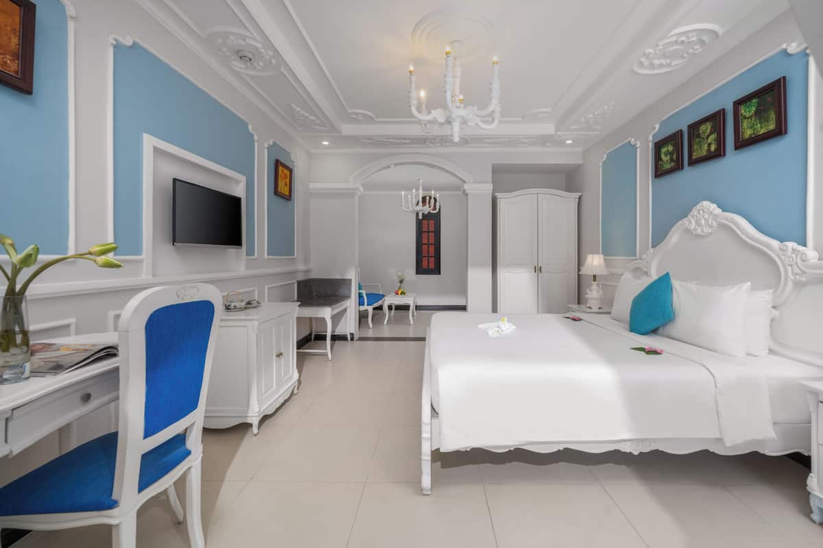 A bedroom with a large bed, a desk, a chair, a television, and a chandelier.