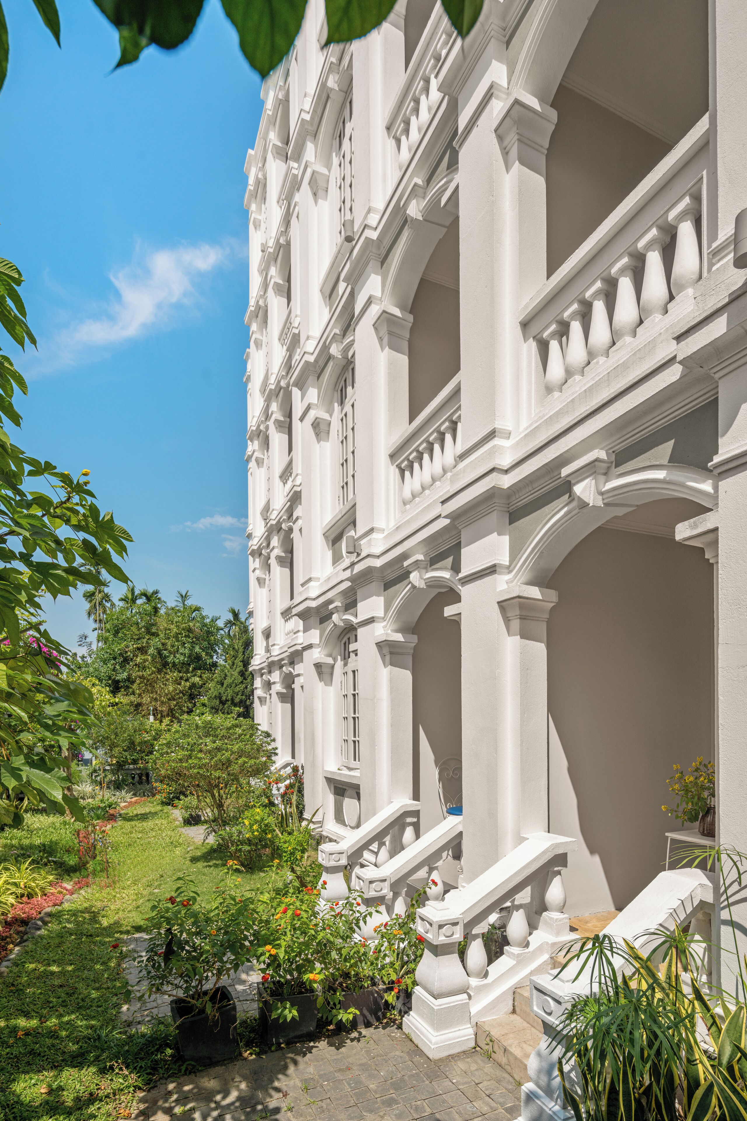 A grand building with classical architecture, featuring multiple columns and a well-maintained garden with various plants and flowers.