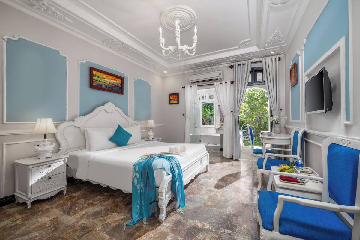 A bedroom with a large bed, a nightstand, a chair, a table with a fruit bowl, and a window with curtains.