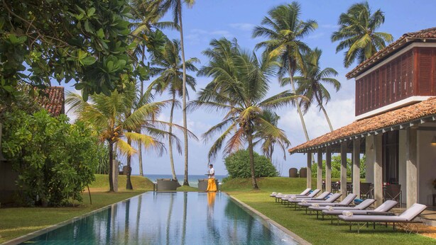 A swimming pool with lounge chairs, palm trees, and a building with a tiled roof. - The Frangipani Tree By Edwards Collection (Unawatuna)