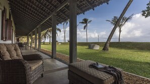 A covered outdoor seating area with wicker furniture and a view of palm trees and a grassy hill. - The Frangipani Tree By Edwards Collection (Unawatuna)